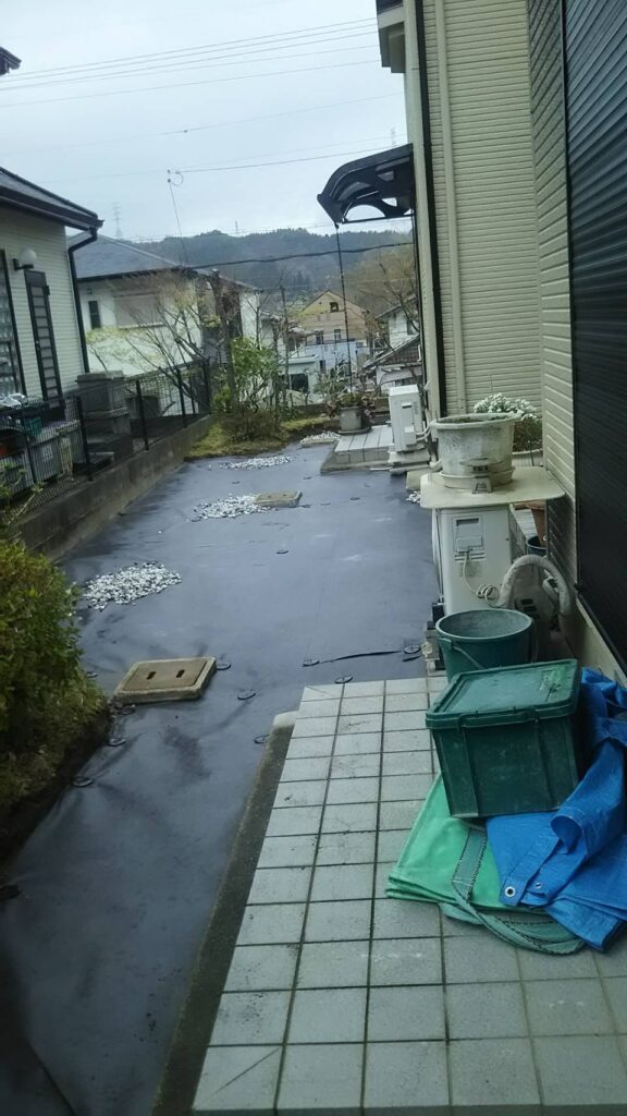 まずは落ち葉や雑草を撤去し、地面を均してから防草シートを全面に敷き詰めます。給水・排水マスのまわりはカットして丁寧に処理し、ピンでしっかりと固定。 その後、五色の化粧砂利を全体にバランスよく敷いていきました。写真でもご覧いただけるように、建物とフェンスの間の細かなスペースまで丁寧に仕上げております。